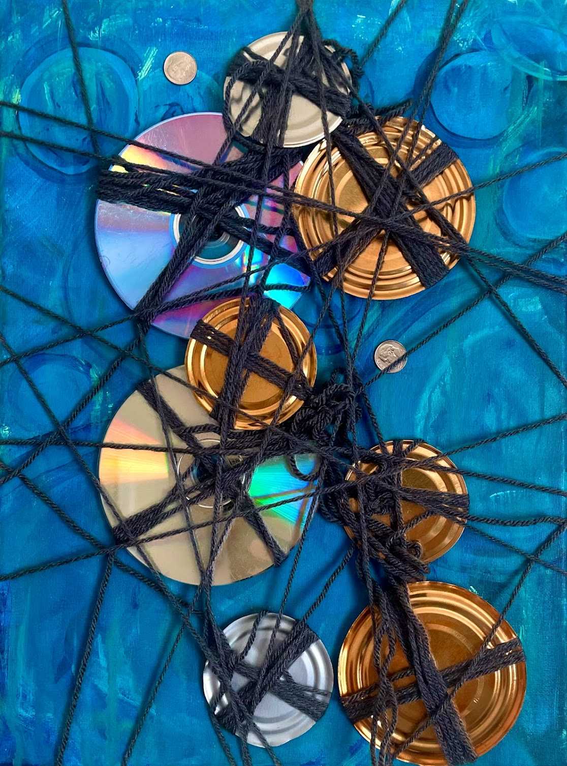 Blue painting, and then with strings attached there is a varied amount of circular objects that are of the likes of the tops of tin cans and cd's neatly arranged, and yarn holding it all together in the center creating a grid like holding structure. 